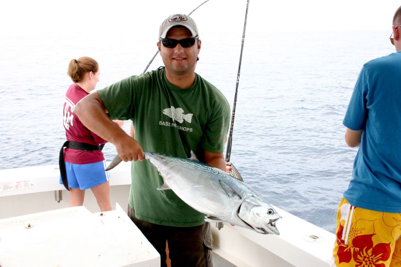 A person in sunglasses proudly holds a large fish on a boat, with others nearby. The ocean is calm under a bright sky, conveying a joyful, relaxed mood.