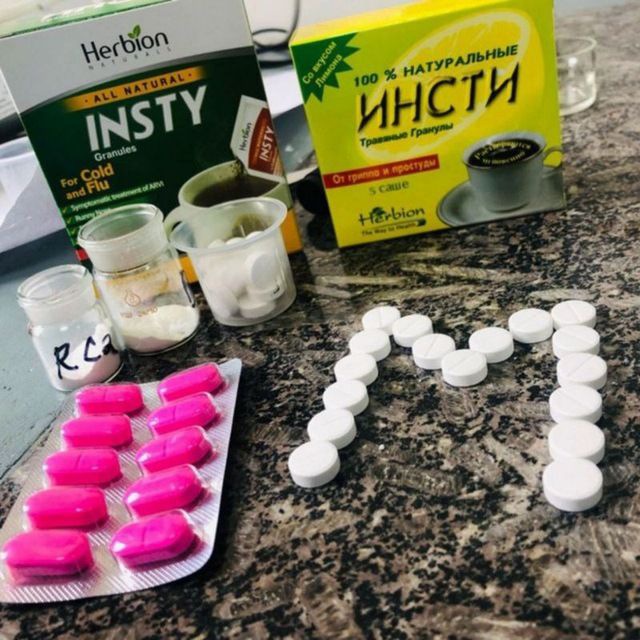 Boxes of herbal cold remedies are on a marble surface. Nearby are pink tablets in a blister pack and white pills arranged to form the letter "M."