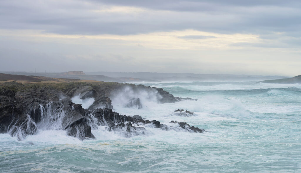 Rocky cliffs meet turbulent ocean waves under a cloudy sky, creating a dramatic and moody seascape. Mist rises from the crashing surf.
