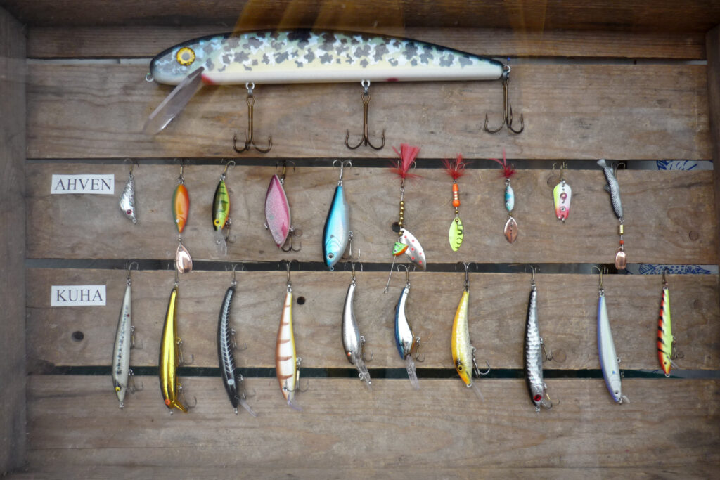 A wooden display showcases two rows of colorful fishing lures labeled "Ahven" and "Kuha." Each lure varies in shape and color, conveying a rustic, crafted aesthetic.