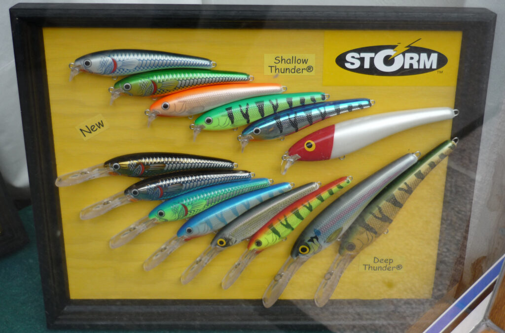 Framed display of colorful fishing lures on a yellow background. Labels "Shallow Thunder" and "Deep Thunder" with a mix of silver, green, and red designs.