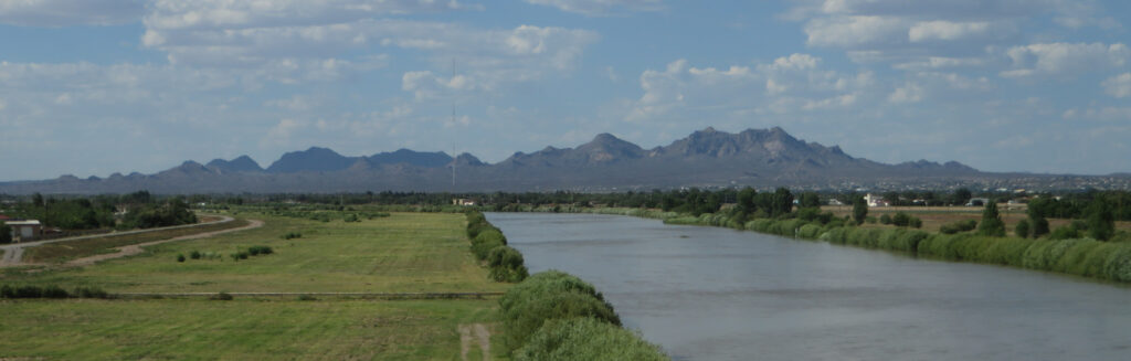 A tranquil river flows through lush green fields with distant mountains under a partly cloudy blue sky, conveying a peaceful, scenic landscape.