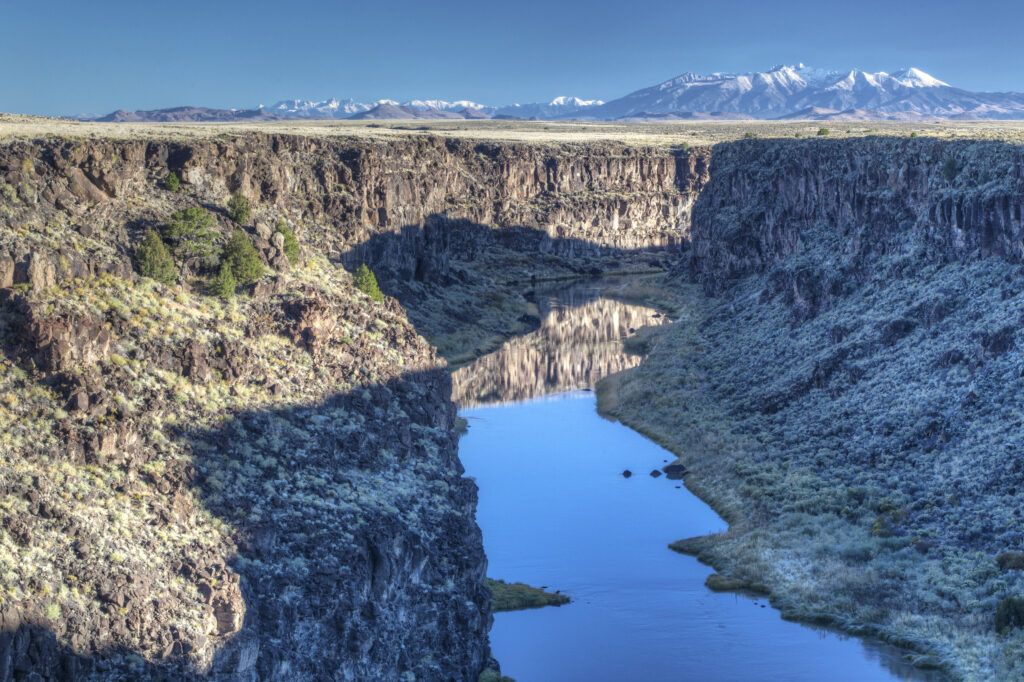 A tranquil river winds through a deep rocky canyon, reflecting the clear blue sky. Snow-capped mountains are visible in the distant background.
