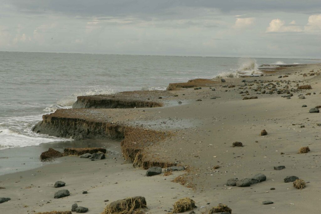 Eroded beach with layers of sand and clay, scattered rocks, and seaweed. Waves crash gently on an overcast day, creating a somber mood.