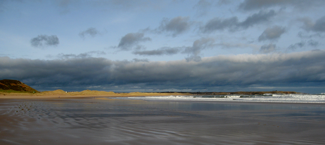 A vast, serene beach with smooth, wet sand under a cloudy blue sky. Gentle waves lap the shore, and distant dunes line the horizon, creating a tranquil scene.