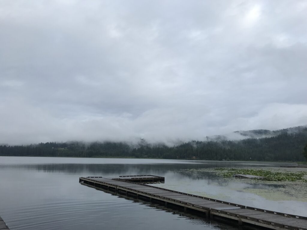 Calm lake scene with a wooden dock extending into the water. Low misty clouds hover over distant forested hills, creating a serene, tranquil atmosphere.