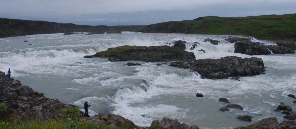 Wide river with cascading waterfalls surrounded by green hills and rocky banks. Two people fish along the shore under an overcast sky.