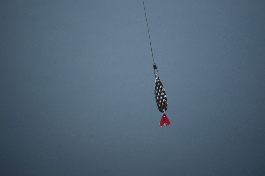 Close-up of a fishing lure with a metallic body and red tail, suspended on a line against a soft blue background, evoking a calm, serene mood.