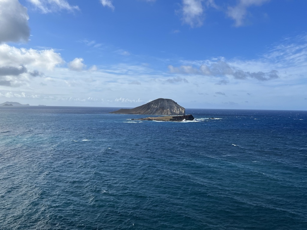 Ocean view with a rocky island in the center, under a bright blue sky with scattered clouds. The sea is deep blue, conveying a calm and serene mood.