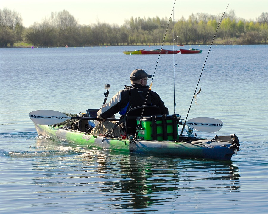 A man kayaks on a calm lake with fishing poles secured to his kayak. He wears a cap and jacket. Trees and colorful kayaks are visible in the background.