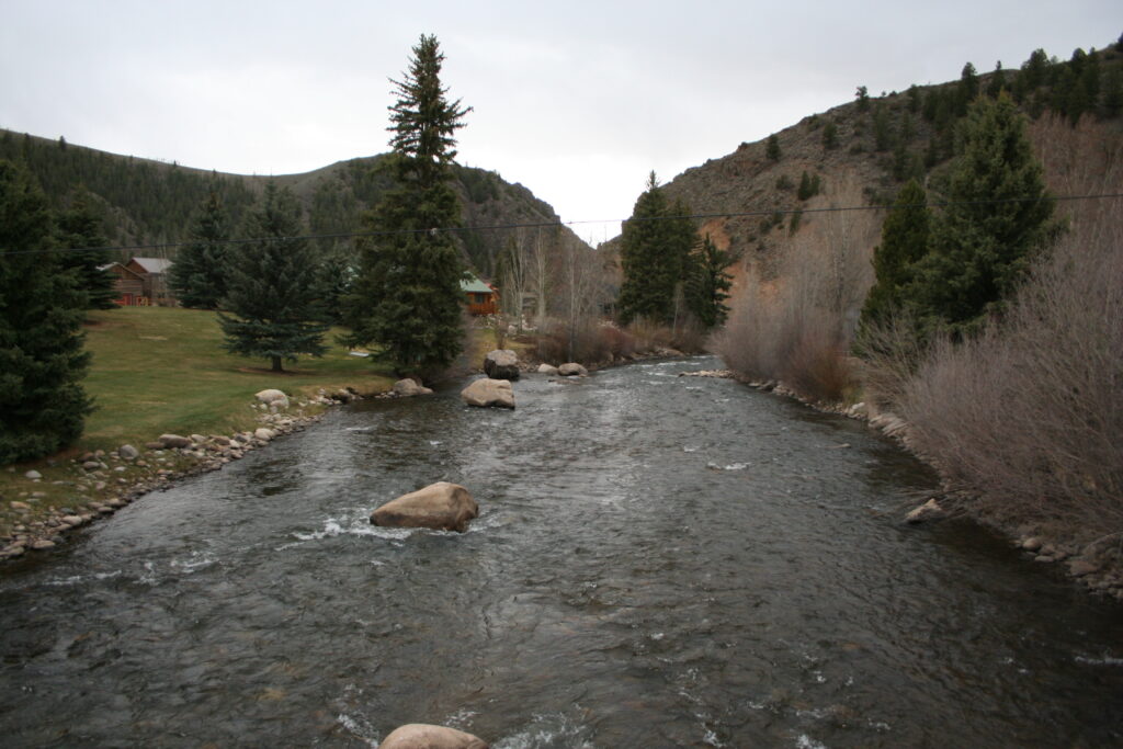 A tranquil river flows through a mountainous landscape, bordered by evergreen trees and rocks. Cabins are nestled among the trees, under an overcast sky.