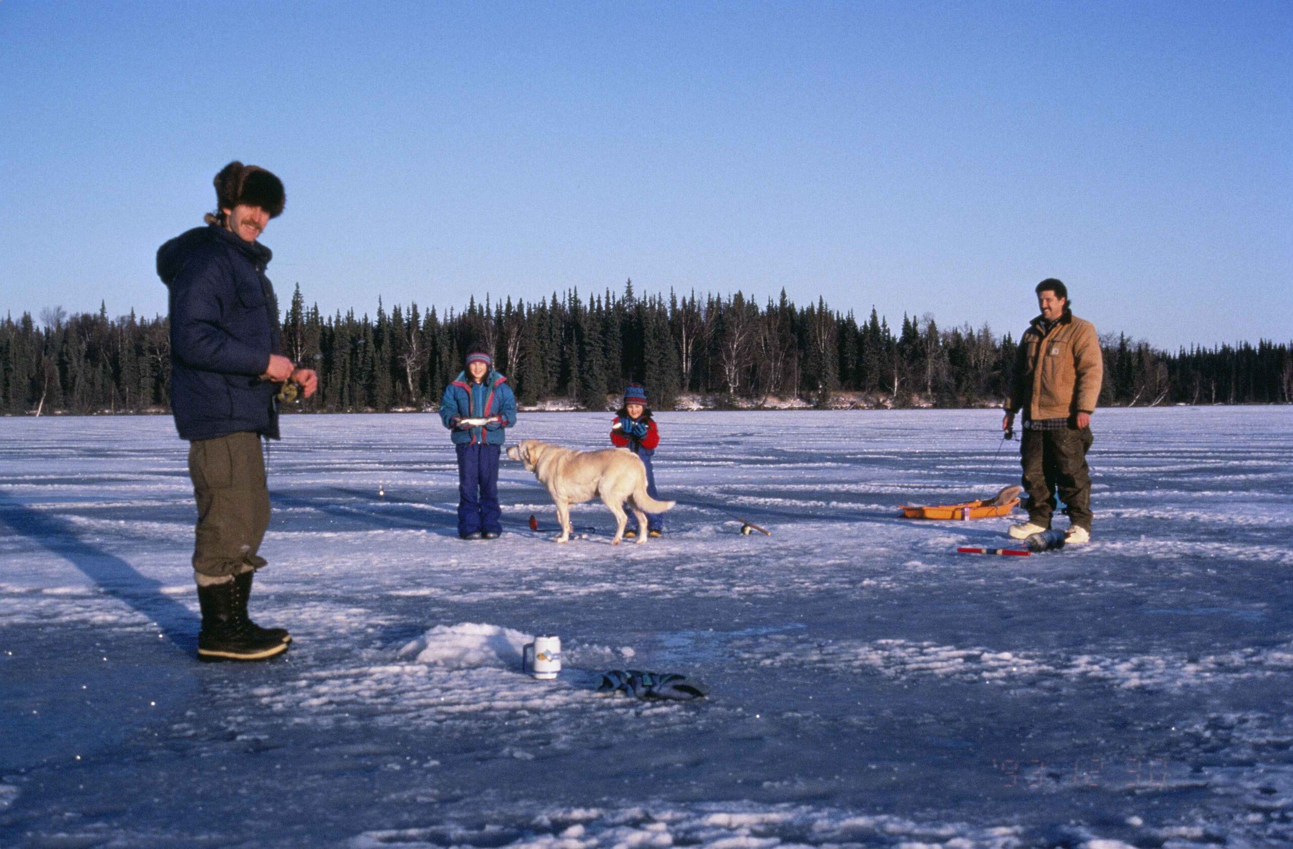 A family ice fishing on a frozen lake under a clear blue sky. Two adults, two children, and a dog stand on the ice, surrounded by snowy equipment.