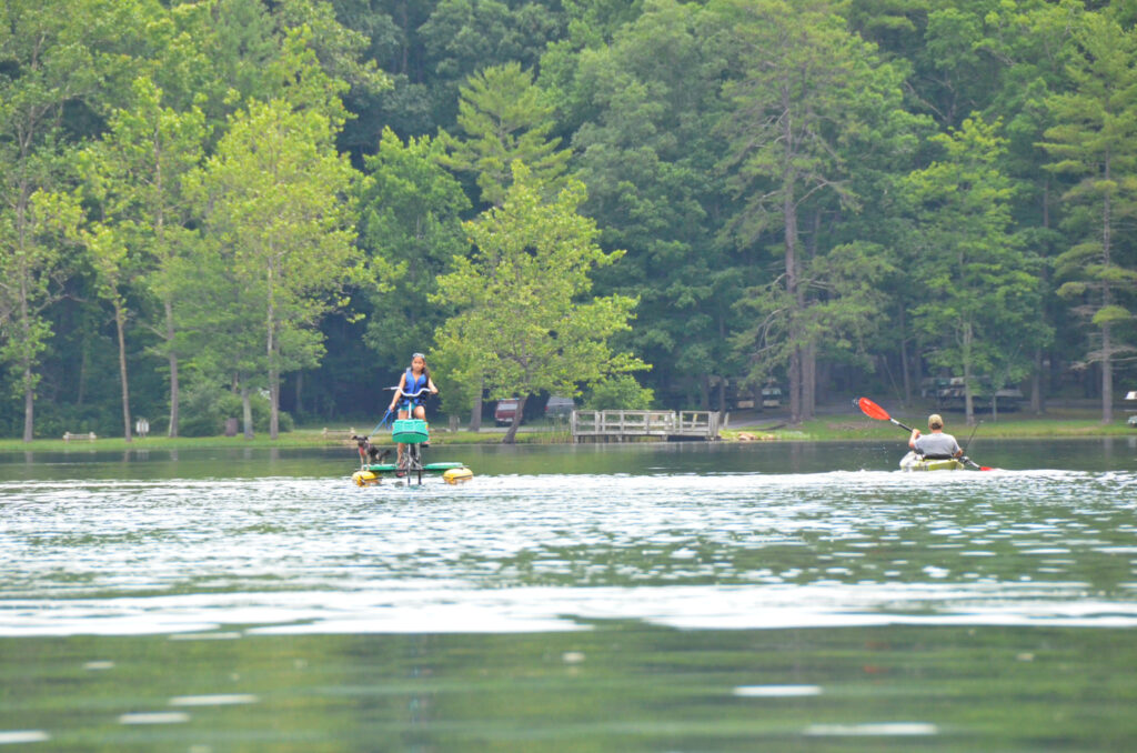 A person rides a water bike with a dog, facing a kayaker with a red paddle on a calm lake surrounded by lush green trees, creating a peaceful scene.