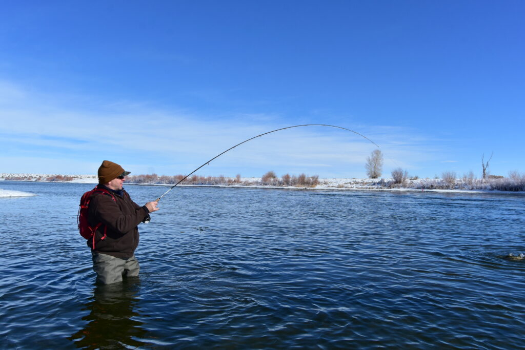 A person stands waist-deep in a wide river, wearing a brown hat and red backpack, fly fishing under a clear blue sky, surrounded by snowy riverbanks.