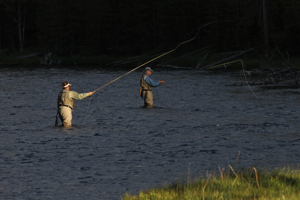 Two people fly-fishing in a river at sunset, dressed in waders and hats. The tranquil scene has a soft, shadowy background of trees and grass.