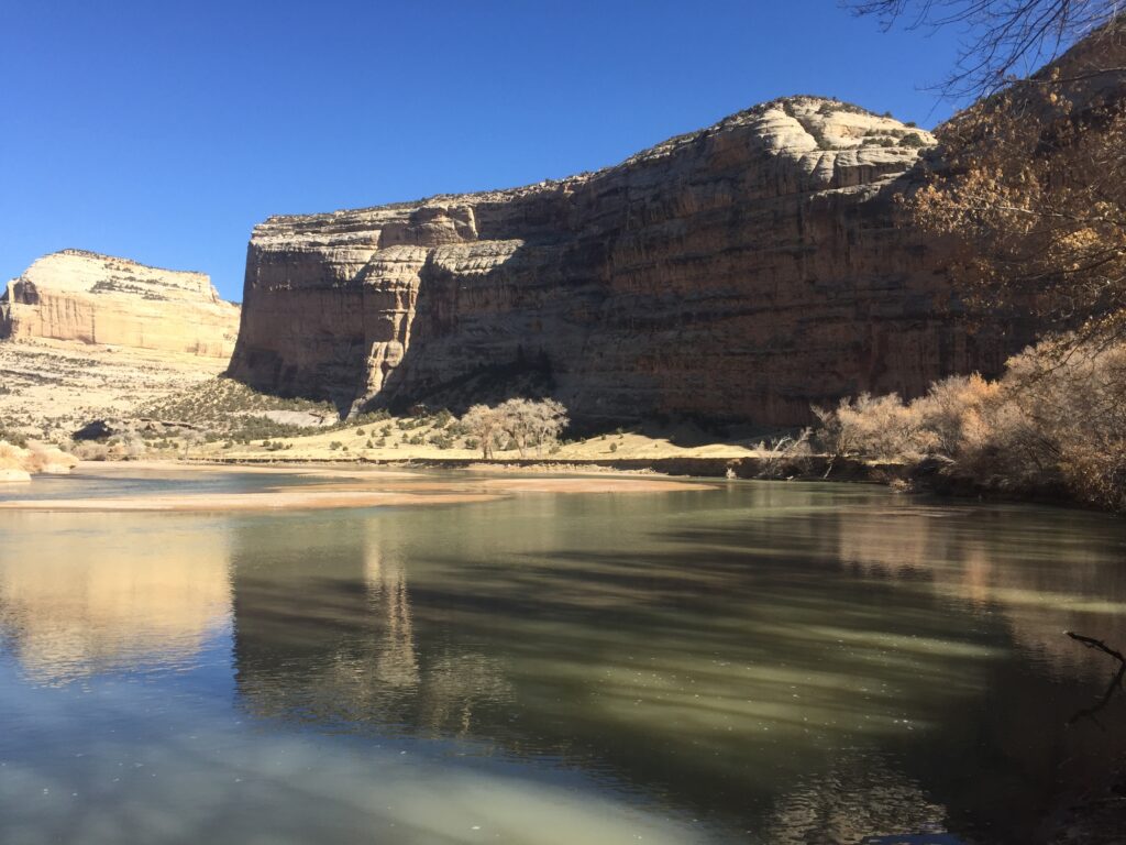 A serene river reflects towering, sunlit cliffs under a clear blue sky. Sparse trees line the bank, basking in the tranquil atmosphere.