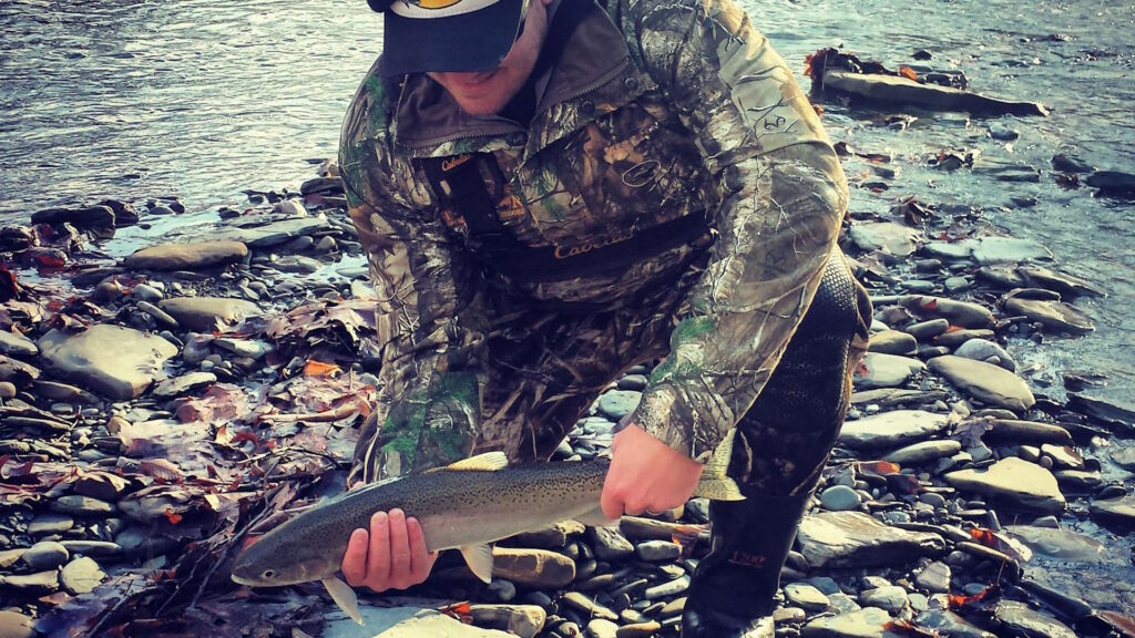 An angler in camouflage gear kneels on a rocky riverbank, holding a freshly caught steelhead trout near a fly fishing rod.