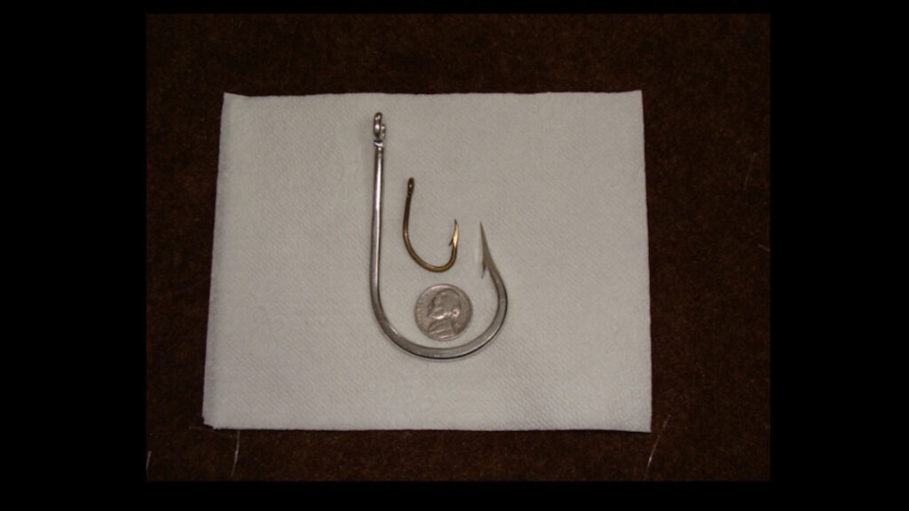 A large silver circle hook, a smaller gold J-hook, and a U.S. dime are displayed on a white napkin for size comparison.