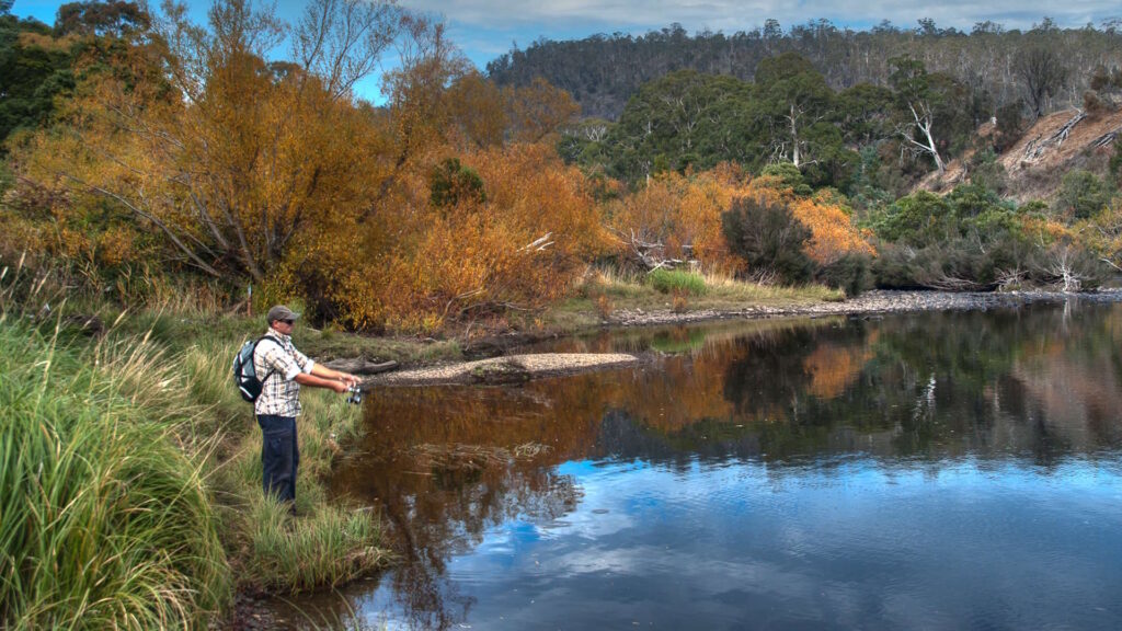 A person fishing in a calm river with autumn-colored trees on the banks.