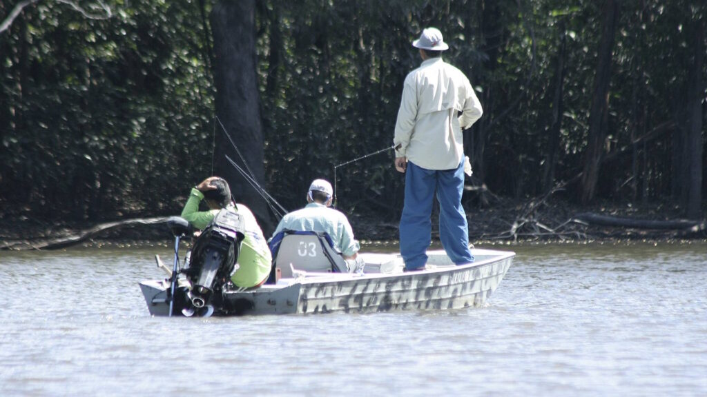 Three people are fishing from a small boat on a calm river