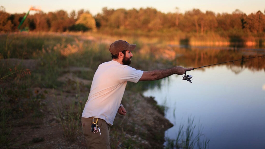 A man in casual attire fishes by a calm lake, reflecting a scenic sunset outdoors
