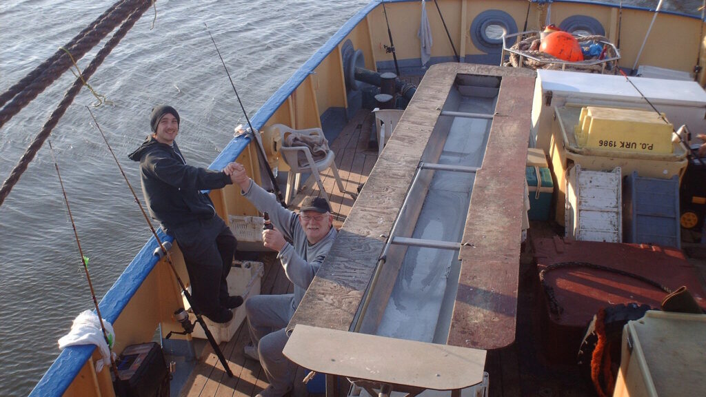 Two men on a boat, one standing and one sitting, holding fishing rods.