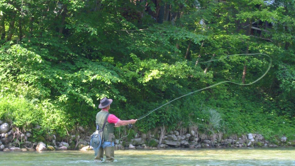 A person in waders and a hat is fly-fishing in a clear river