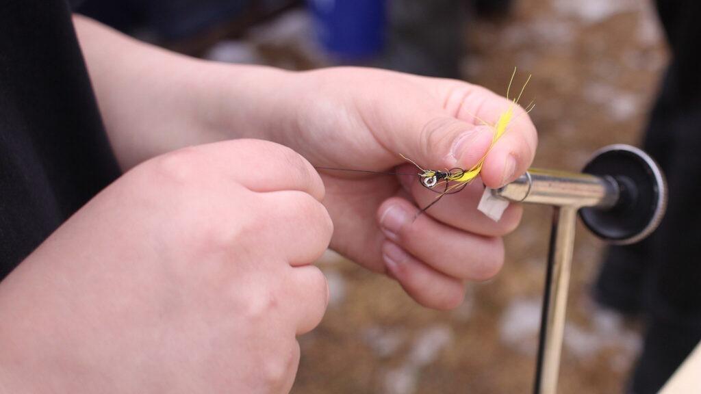 Close-up of hands tying a yellow fly on a hook using thread and a fly-tying vise.