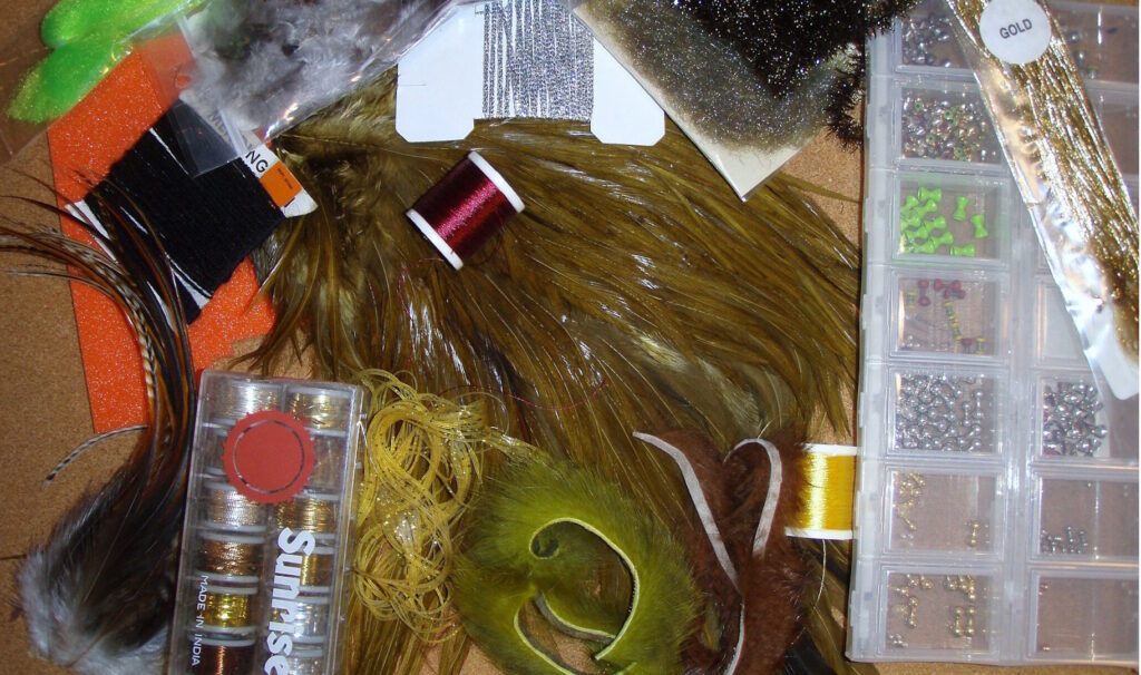 An assortment of fly tying materials including feathers, threads, chenille, hooks, beads, foam, and synthetic fibers.