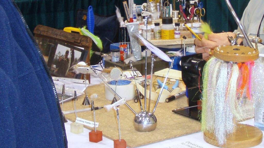 A cluttered fly tying workstation with various tools, feathers, threads, and materials organized for fly creation.
