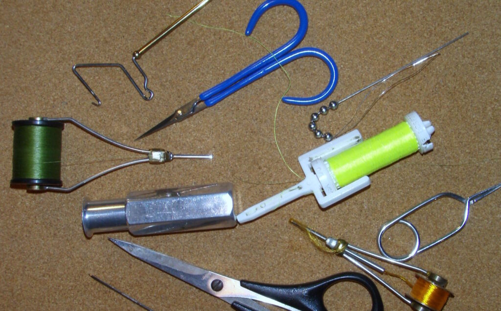A collection of fly tying tools including scissors, bobbins, thread spools, a whip finisher, bodkin, and hackle pliers arranged on a cork surface.