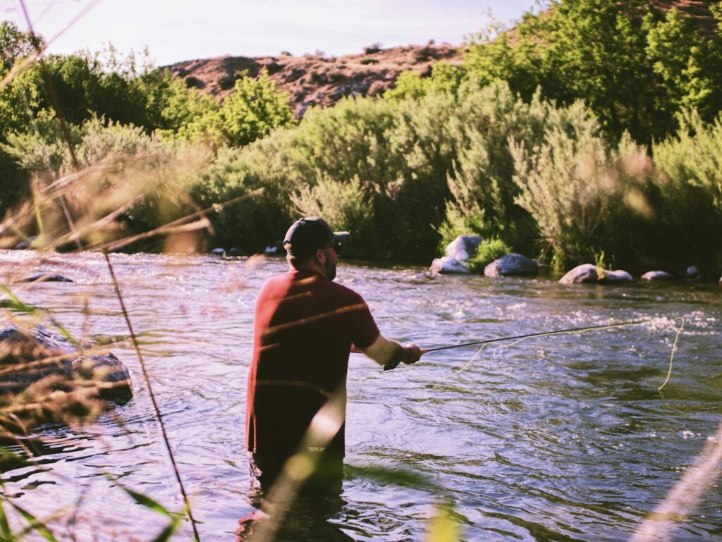 Man in red shirt and black shorts wading in the water, fishing.