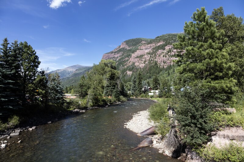 Mountain landscape with a clear river flowing amid lush green trees. A rocky mountain looms in the background under a bright blue sky.
