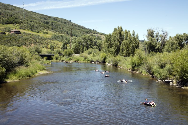People float leisurely on green inner tubes down a tranquil, tree-lined river under a bright blue sky, surrounded by lush hills and greenery.