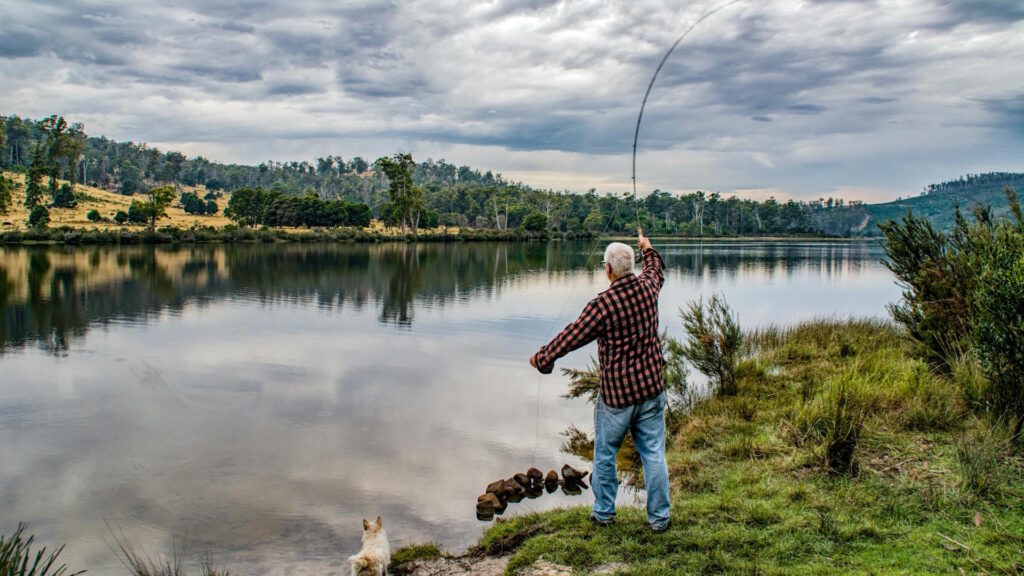 Elderly man fishing by a picturesque lake in Ouse, Tasmania, with a dog by his side.