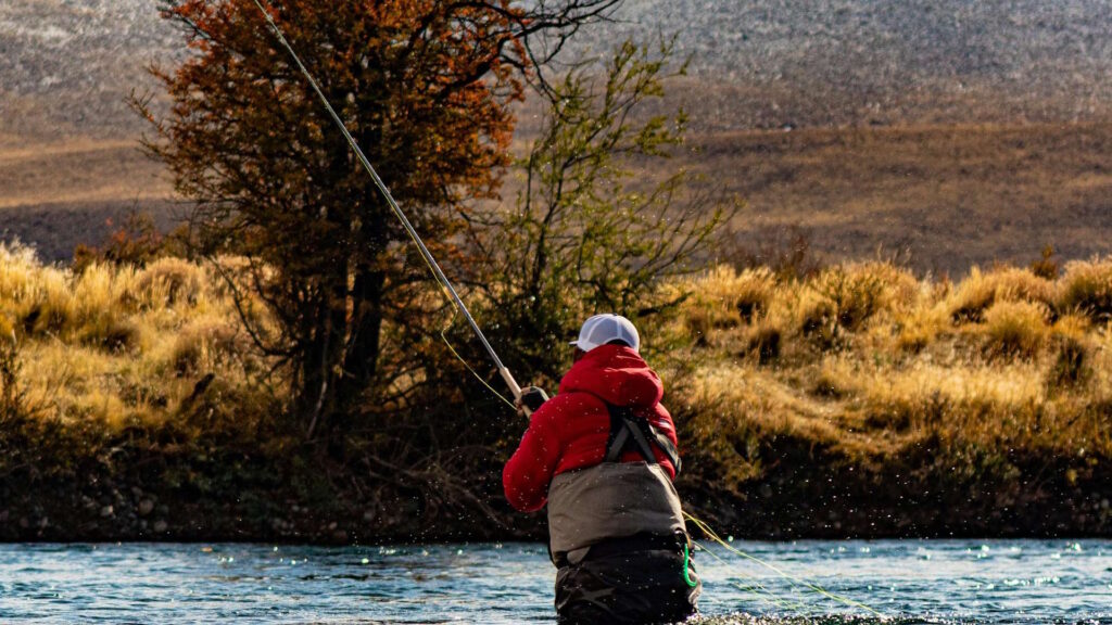 A person in waders and a red jacket fly fishing in a river during autumn, with trees and dry grass on the banks.