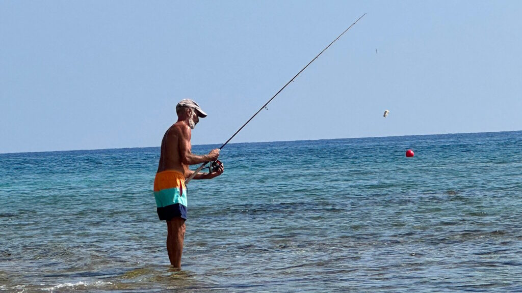 A man in colorful swim trunks stands in shallow sea water, casting a fishing line with a spinning rod.