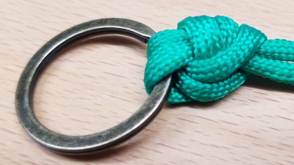 Close-up of a green rope tied with a Palomar knot to a metal ring on a wooden surface.
