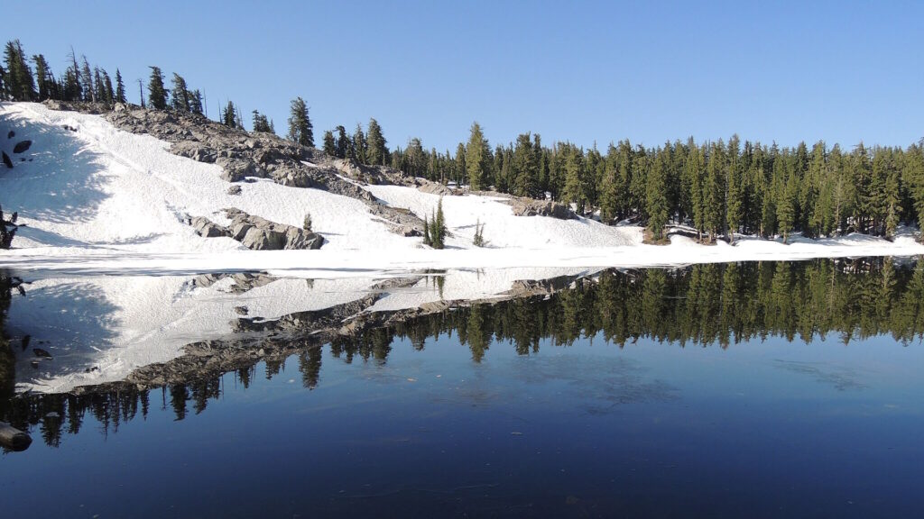 A clear alpine lake reflects snow-covered slopes and dense evergreen forest under a bright blue sky.