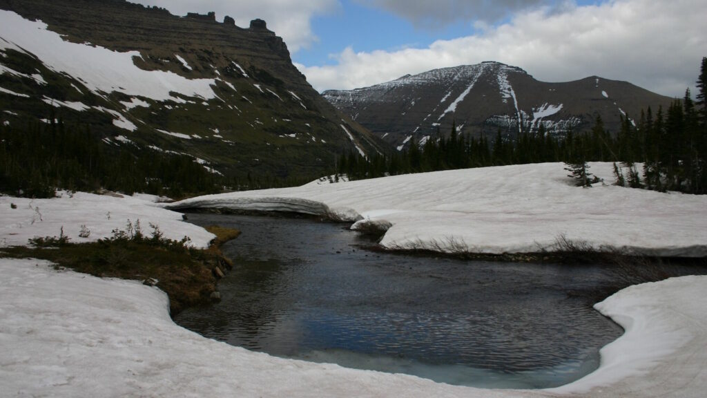 Snow-covered mountain landscape with a small river emerging from melting snowpack in a high-altitude valley.