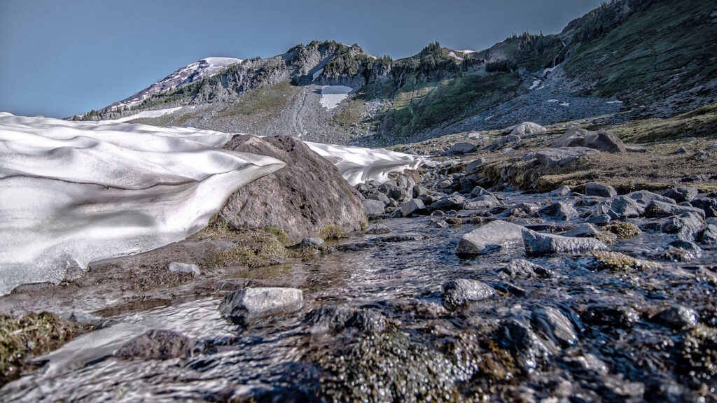 A stream flows through a rocky, melting snowfield with mountains in the background under a clear sky