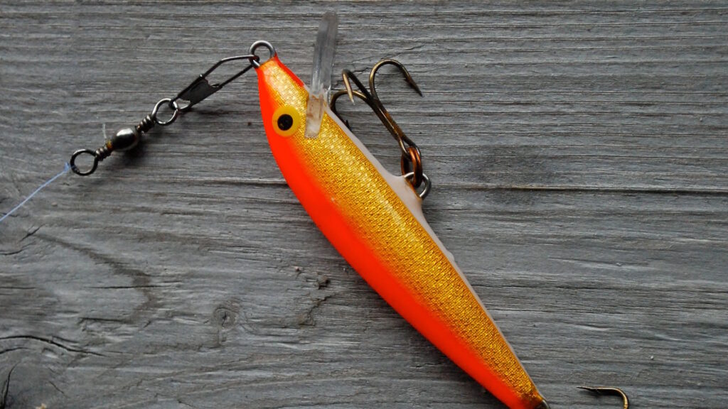 A brightly colored orange and yellow fishing lure with treble hooks lying on a wooden surface.