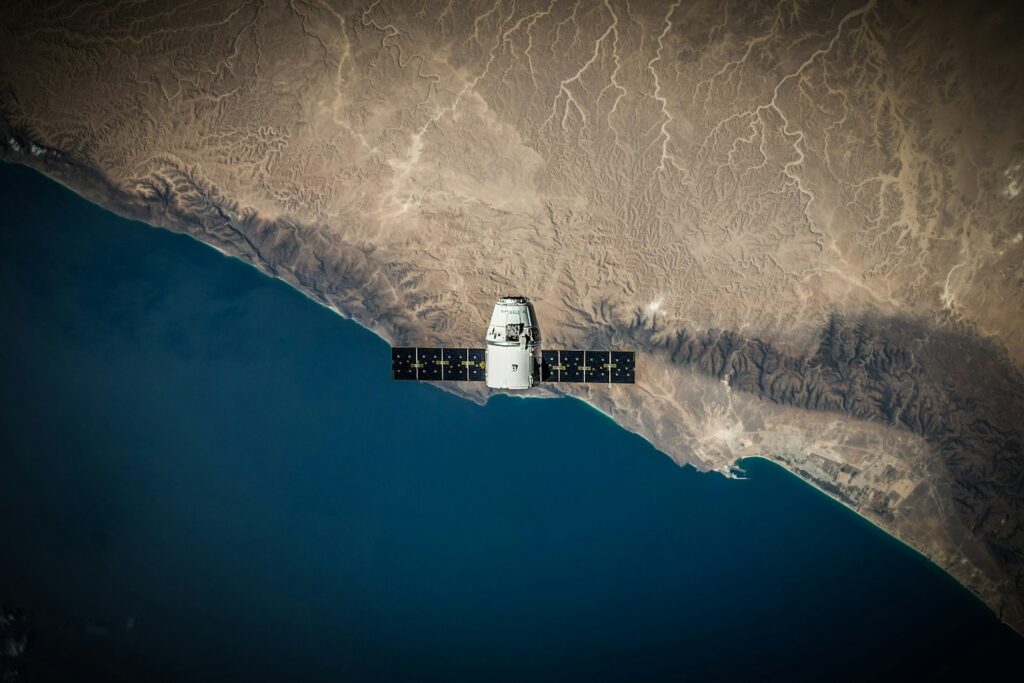 A satellite hovers above a rugged coastline, with desert landscapes meeting deep blue ocean. It conveys technological precision and Earth's vastness.