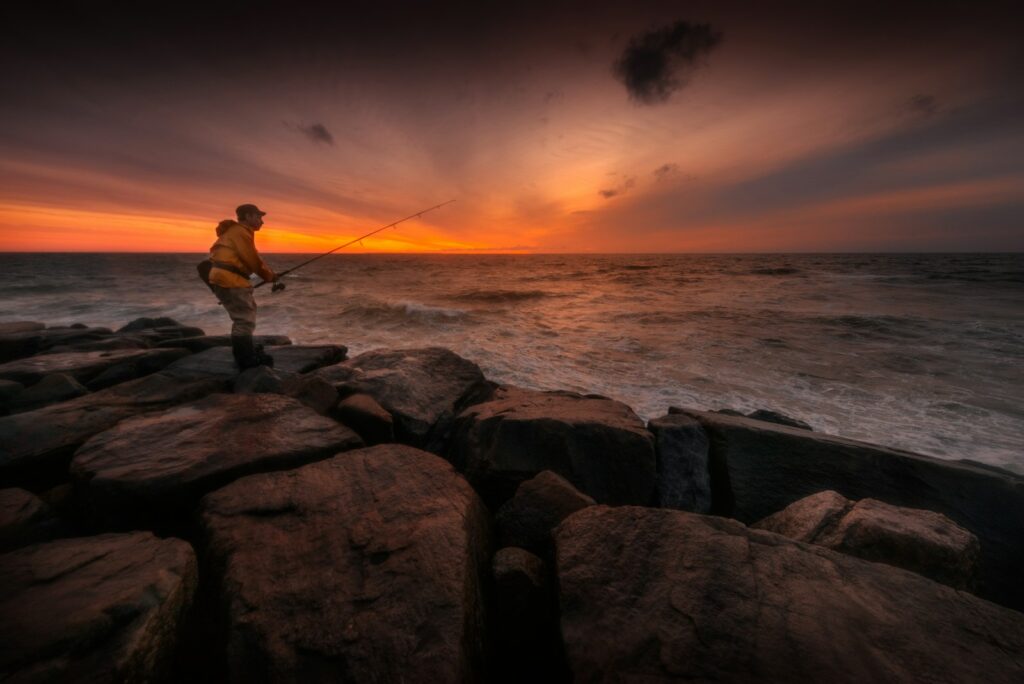 A man standing on rock fishing on body of water during sunset.