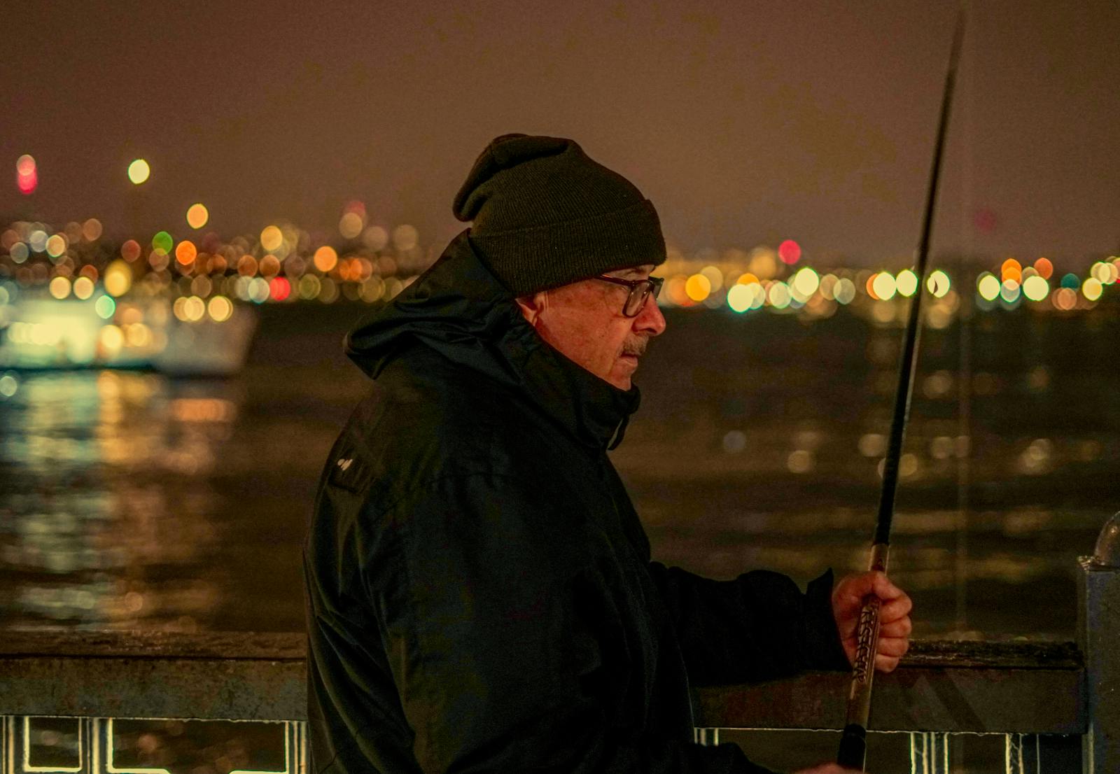 Elderly man fishing at night by the waterfront, city lights in background.