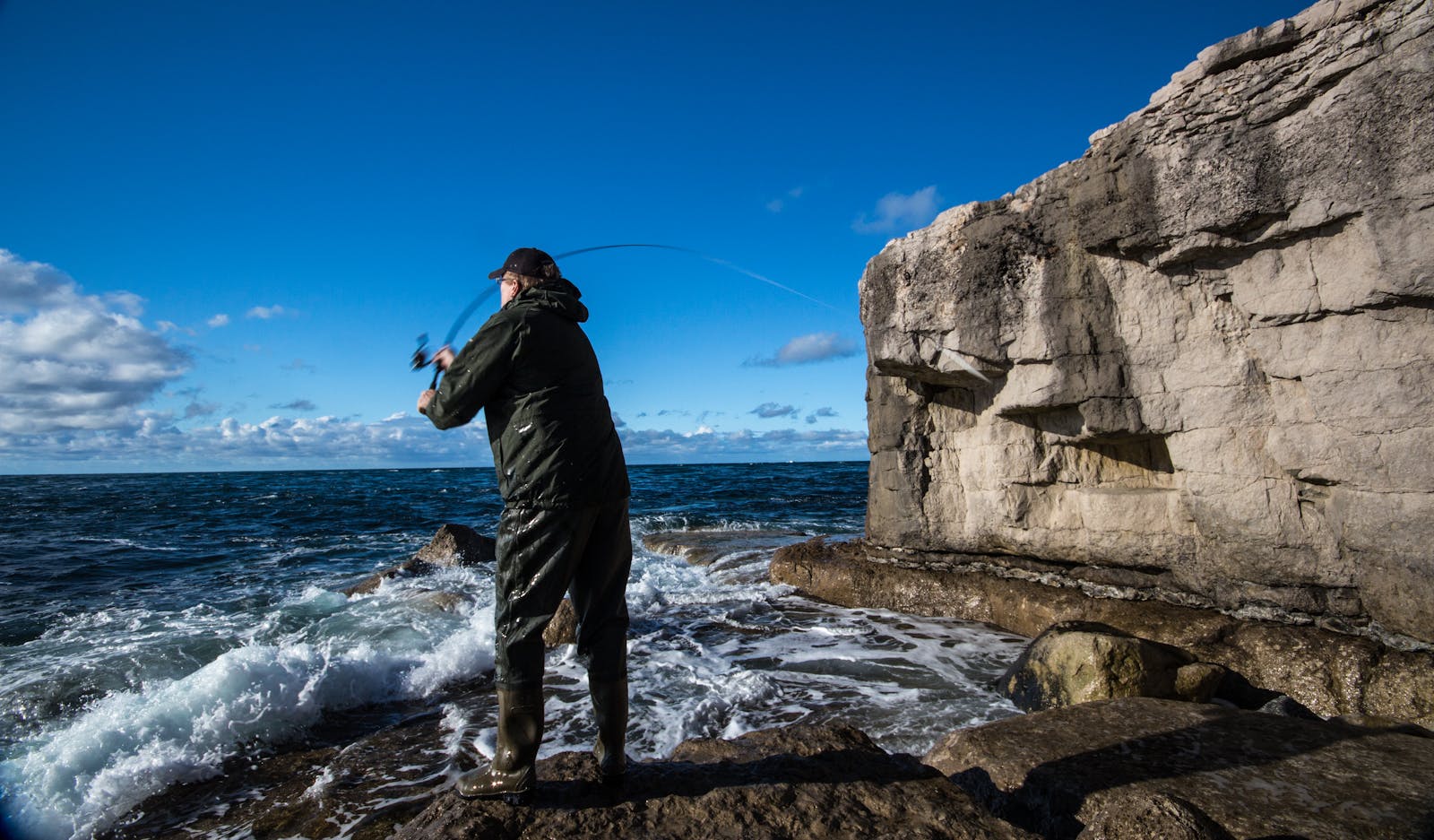 Angler casting line by rocky cliffs at England's scenic Jurassic Coast under vibrant blue skies.