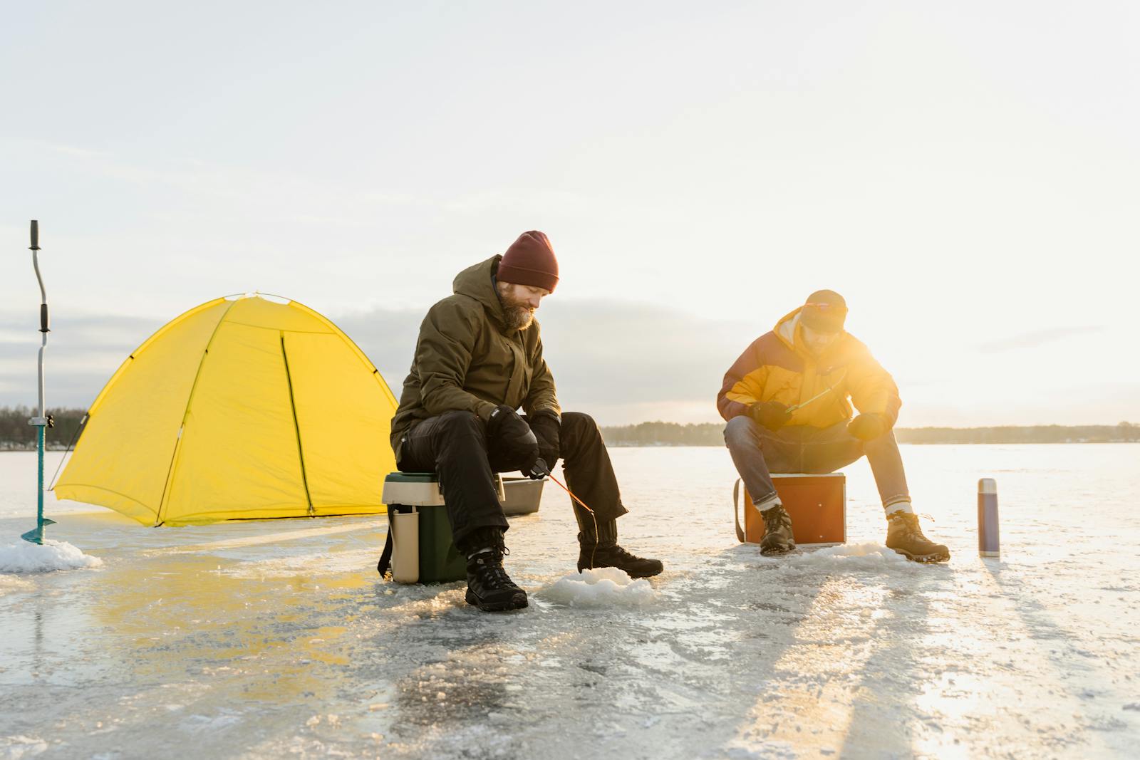 Two men enjoying ice fishing on a frozen lake beside a bright yellow tent.