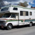A vintage white Winnebago RV is parked on a sunny residential street. It features brown and green stripes and rectangular windows, conveying a nostalgic, adventurous tone.