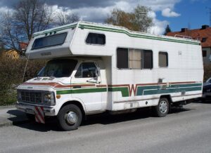 A vintage white Winnebago RV is parked on a sunny residential street. It features brown and green stripes and rectangular windows, conveying a nostalgic, adventurous tone.