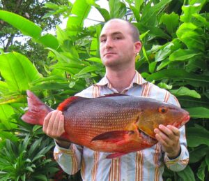A man in a striped shirt holds a large red snapper fish, standing in front of lush green tropical foliage. He has a proud expression.